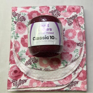 Puede incluir: Una madeja de hilo de crochet Aunt Lydia's Classic 10 de color burdeos oscuro. El hilo es 100% algod&oacute;n y se muestra sobre un fondo blanco con un patr&oacute;n floral rosa.