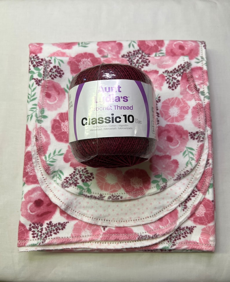 Puede incluir: Una madeja de hilo de crochet Aunt Lydia's Classic 10 de color burdeos oscuro. El hilo es 100% algod&oacute;n y se muestra sobre un fondo blanco con un patr&oacute;n floral rosa.