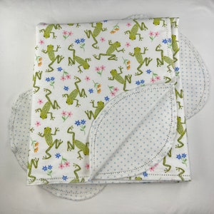 May include: A white baby blanket with a green frog and flower pattern on one side and a white background with blue polka dots on the other side.