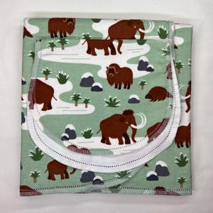 May include: A folded baby blanket with a light green background and a repeating pattern of brown mammoths, trees, and snowy hills. The blanket has a white border with a dotted trim.