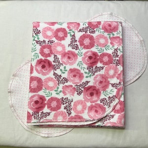 Puede incluir: Una manta de beb&eacute; de color rosa y blanco con estampado floral y borde festoneado. La manta est&aacute; doblada por la mitad y se encuentra sobre una superficie blanca.