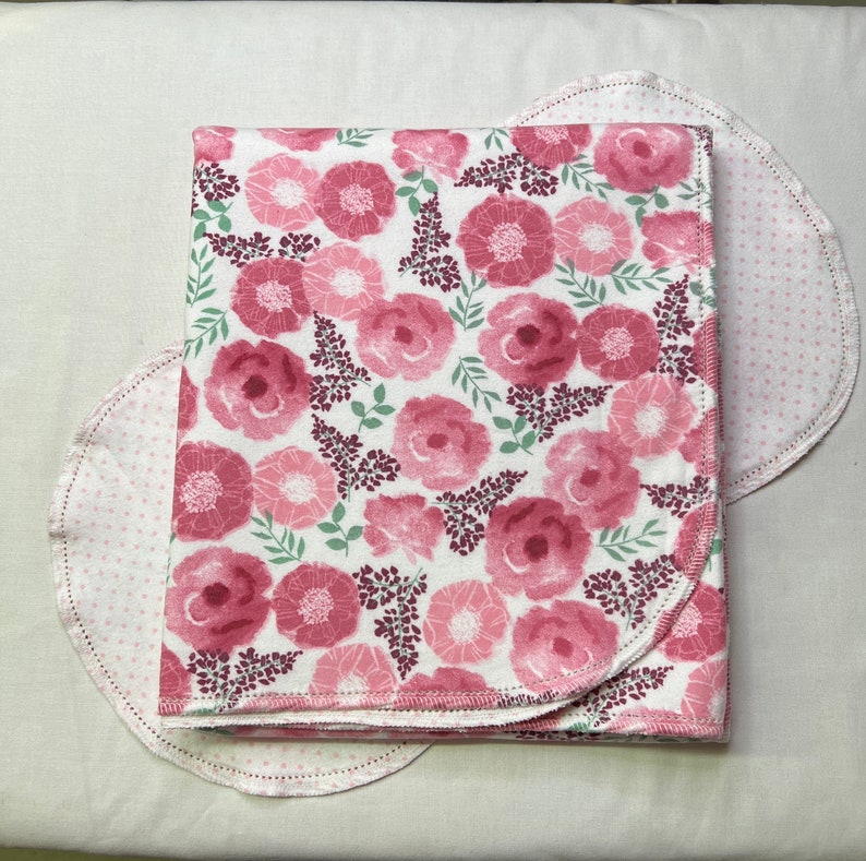 Puede incluir: Una manta de beb&eacute; de color rosa y blanco con estampado floral y borde festoneado. La manta est&aacute; doblada por la mitad y se encuentra sobre una superficie blanca.