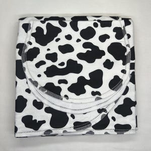 May include: Folded baby blanket with a black and white cow print. The blanket has a rounded edge and is made of soft fabric. The cow print features irregular black spots on a white background. The blanket is folded neatly.