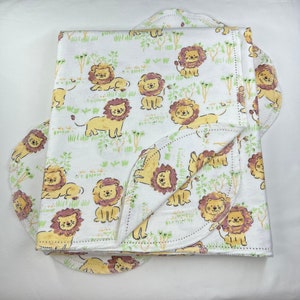 May include: A white baby blanket with a yellow lion print. The blanket is folded in half and has a scalloped edge.