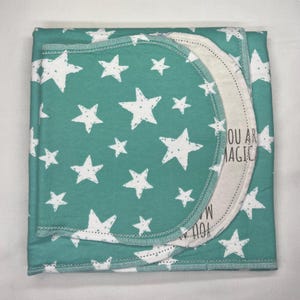 May include: A folded teal blanket with white star patterns. A circular section with the text "YOU ARE MAGIC" is visible. The blanket is made of soft material and is suitable for babies or children.