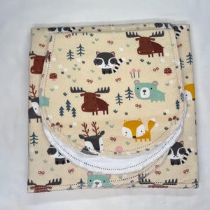May include: A folded baby blanket with a cream-colored background and a repeating pattern of woodland animals, including moose, raccoons, foxes, and bears. The blanket has a white, curved edge and is decorated with trees and mushrooms.