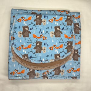 May include: A baby blanket and burp cloth set with a light blue background and a repeating pattern of brown bears, orange foxes, and white mountains. The burp cloth has a brown border and is folded over the blanket.