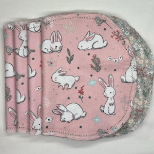 May include: A stack of pink, oval-shaped baby cloths with a white bunny and floral pattern. The edges are stitched with a pink thread. The cloths are made of soft fabric.