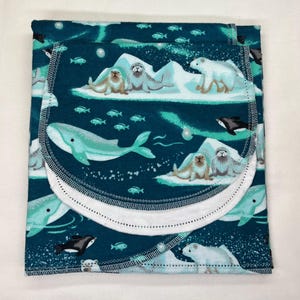 May include: A teal baby blanket with a marine life design. The blanket features illustrations of whales, seals, polar bears, and fish. The edges are trimmed with white stitching. The overall design has a whimsical, aquatic theme.