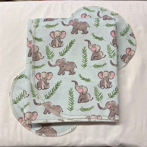 May include: A light blue baby blanket and burp cloth set. The blanket and burp cloth feature a repeating pattern of gray elephants and green fern fronds. The edges of the blanket and burp cloth are finished with a white stitch.
