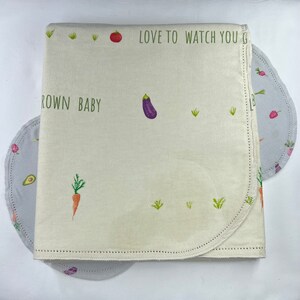 May include: Cream-colored baby blanket with a vegetable theme. The blanket features illustrations of carrots, an eggplant, and a tomato, along with the text "LOVE TO WATCH YOU GROW BABY". Two gray circular pads with vegetable illustrations are also visible.
