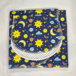 May include: A folded, navy blue baby blanket with a celestial pattern. The design features smiling yellow suns, moons, stars, planets, and rainbows. The blanket has a white, dotted trim and a matching bib.