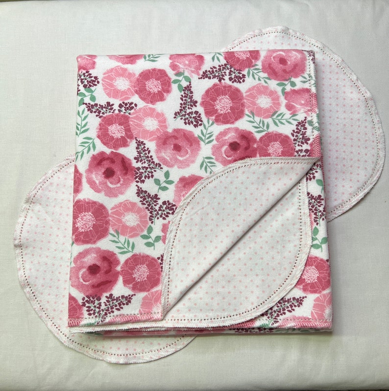 Puede incluir: Una manta de beb&eacute; rosa y blanca con estampado floral y un reverso blanco con lunares. La manta est&aacute; doblada por la mitad y tiene un borde festoneado.
