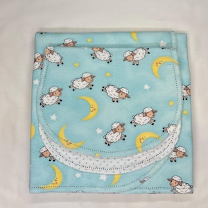 May include: A light blue baby blanket and burp cloth set. The set features a repeating pattern of white sheep, yellow crescent moons, and stars. The burp cloth is layered with a white dotted trim.
