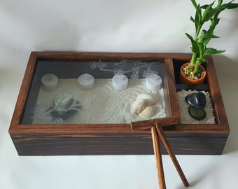May include: A wooden zen garden with a glass top, featuring white sand, small candles, decorative shells, and a small bamboo plant. The dark brown frame holds the components, creating a miniature, tranquil landscape. Includes a wooden rake.