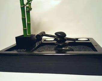 May include: Black wooden tray with a small potted bamboo plant, a rake, a pile of black stones, and two white tea lights.