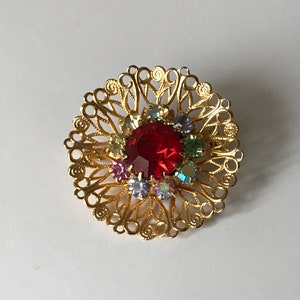 May include: A gold-toned brooch with a central red gemstone surrounded by smaller, colorful gems. The brooch features an openwork design with intricate, swirling patterns. The overall shape is circular, and the brooch appears to be vintage.