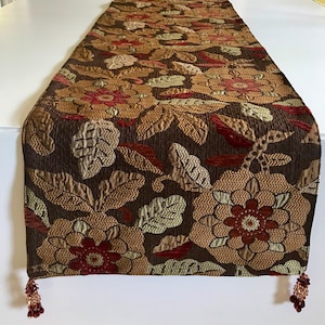 May include: A rectangular table runner featuring a dark brown background with a woven floral pattern in shades of gold, tan, and deep red. The fabric has a textured surface and is finished with small, beaded tassels at each corner of the end.