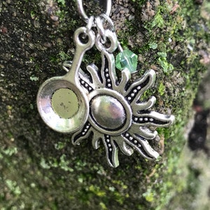 May include: Silver necklace with a sun charm and a small spoon charm. A green crystal bead hangs from the sun charm.