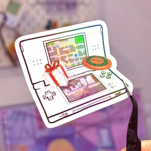May include: A die-cut sticker featuring a white handheld gaming console with a dual-screen display. The top screen shows a colorful map, while the bottom screen displays a dog. A small gift box with a red ribbon and an orange disc are on the console.