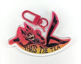 May include: An acrylic keychain featuring a cartoon character with red hair, a sharp-toothed grin, and a red suit, holding a teacup. The character is positioned above a yellow and orange gradient banner with the black text THAT'S THE TEA.