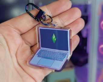 Puede incluir: Un llavero de acrílico transparente con forma de portátil, con marco morado y pantalla azul oscuro que muestra un icono de plumbob verde brillante en forma de diamante. El llavero está unido a un cierre de mosquetón metálico iridiscente.