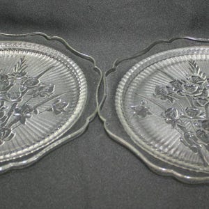 May include: Two clear glass plates featuring a pressed floral design of a bouquet of roses and leaves in the center. The plates have a circular shape with a scalloped, wavy edge and a textured, radiating sunburst pattern surrounding the flowers.