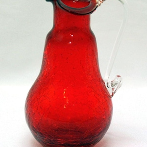 May include: A vibrant red glass pitcher featuring a distinct crackle texture throughout the body. The vessel has a pear-shaped silhouette, a wide pouring spout with a ruffled rim, and a smooth, clear glass handle attached to the side.