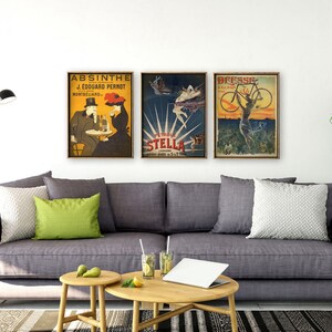 Puede incluir: Tres carteles vintage enmarcados en madera, colgados en una pared blanca sobre un sof&aacute; gris. Los carteles son para Absenta, Stella y Bresse. El sof&aacute; tiene cojines verdes y blancos y un ordenador port&aacute;til sobre una mesa de caf&eacute; de madera delante de &eacute;l.