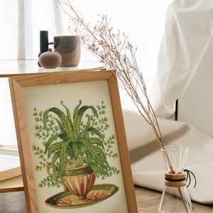 May include: A framed botanical print of a fern plant growing in a teacup. The print is in a light wood frame and is sitting on a table with a vase of dried flowers.