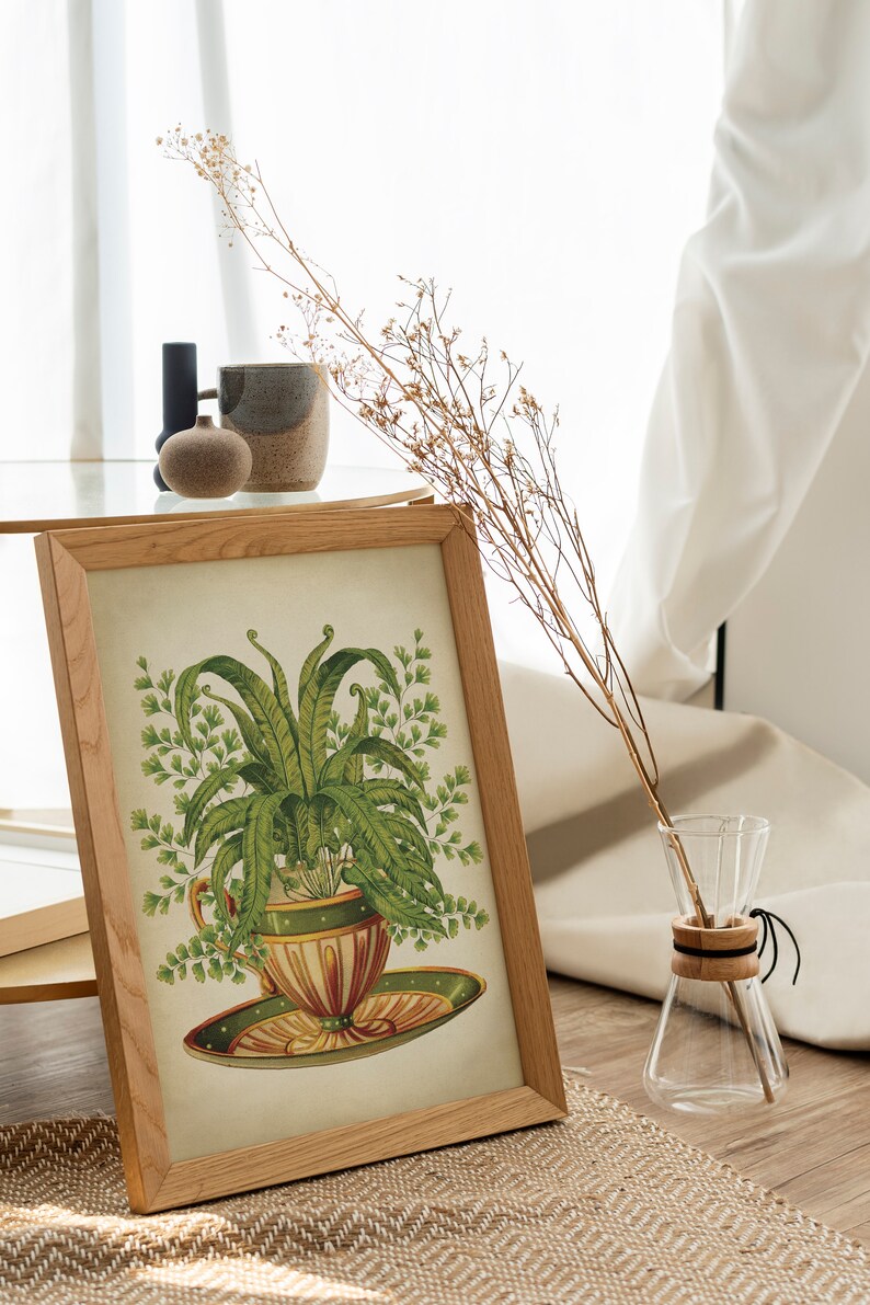 May include: A framed botanical print of a fern plant growing in a teacup. The print is in a light wood frame and is sitting on a table with a vase of dried flowers.
