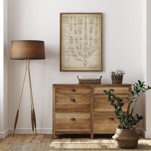 May include: A vintage botanical illustration of a tree with branches and leaves, printed on a beige background and framed in a brown wood frame. The print is hanging on a white wall above a wooden dresser with six drawers. A potted plant and a basket of books are on top of the dresser.