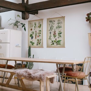 May include: A wooden dining table with a light wood finish and a white background. The table is set with four chairs, two of which are gold metal chairs with brown cushions. There are two botanical prints hanging on the wall behind the table.