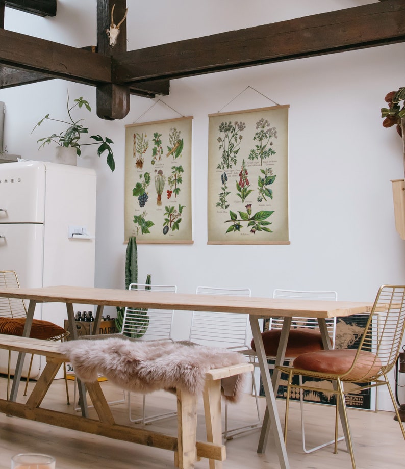 May include: A wooden dining table with a light wood finish and a white background. The table is set with four chairs, two of which are gold metal chairs with brown cushions. There are two botanical prints hanging on the wall behind the table.