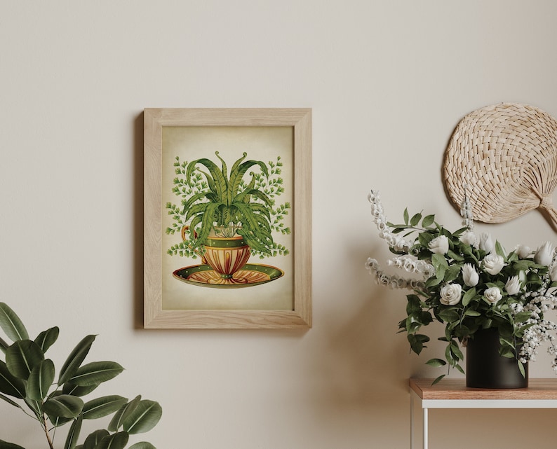 May include: A vintage illustration of a fern plant growing in a teacup and saucer. The teacup is decorated with green and gold accents. The illustration is framed in a light wood frame.