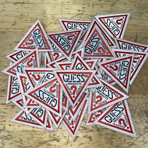 Può includere: Una collezione di toppe triangolari ricamate con bordo rosso e sfondo bianco. Ogni toppa mostra il testo nero GUESS PRODUCTS, la parola WASHED e un logo con punto interrogativo rosso centrato all'interno della forma triangolare.