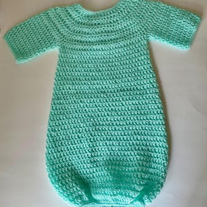 May include: A mint green, handmade crochet baby sleep sack. The sleep sack has short sleeves and a rounded neckline. The crochet stitch creates a textured appearance. The sleep sack is designed to keep a baby warm and comfortable.