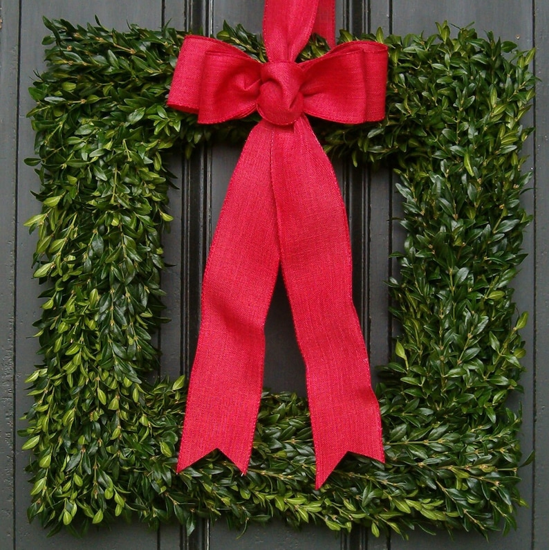 May include: A red bow with a large, square boxwood wreath. The wreath is made of green boxwood leaves and is perfect for a front door.