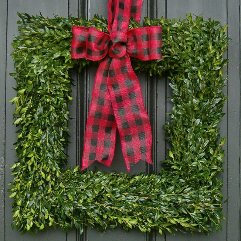 May include: A square green boxwood wreath with a red and black buffalo plaid bow. The wreath is made of boxwood and is perfect for a farmhouse or rustic decor style.