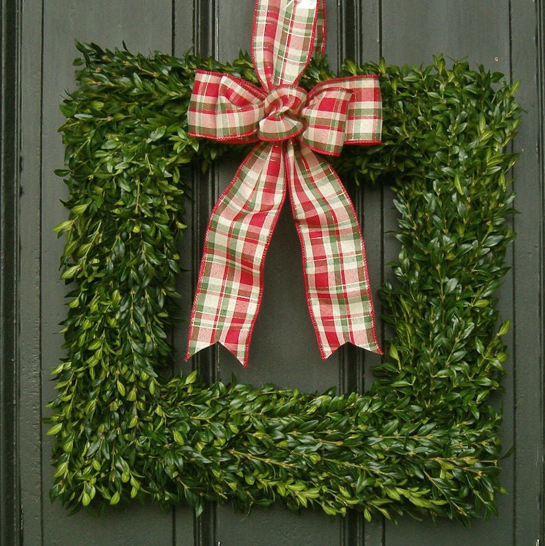 May include: A green boxwood wreath with a red and white plaid bow. The wreath is hanging on a dark wooden door.