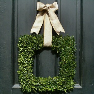 May include: A square boxwood wreath with a beige bow tied on top. The wreath is hanging on a dark green door.