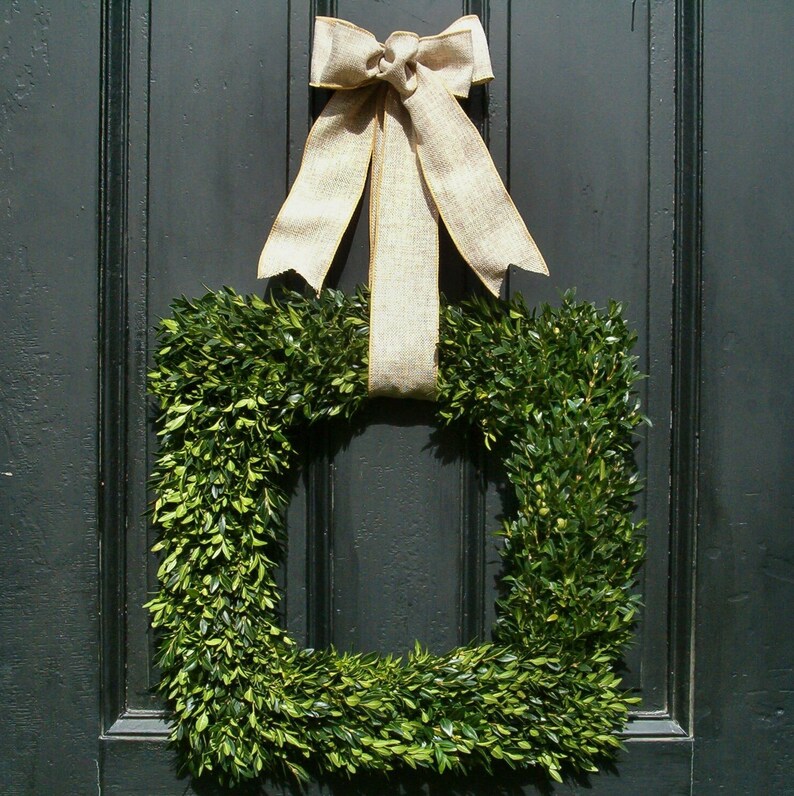 May include: A square boxwood wreath with a beige bow tied on top. The wreath is hanging on a dark green door.