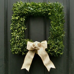 May include: A green boxwood wreath with a large, beige bow. The wreath is hanging on a dark wooden door.