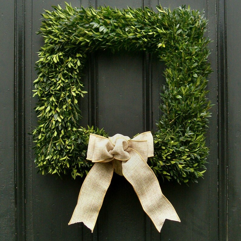May include: A green boxwood wreath with a large, beige bow. The wreath is hanging on a dark wooden door.