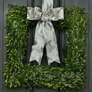 May include: A square boxwood wreath with a large silver satin bow. The wreath is hung on a black door.