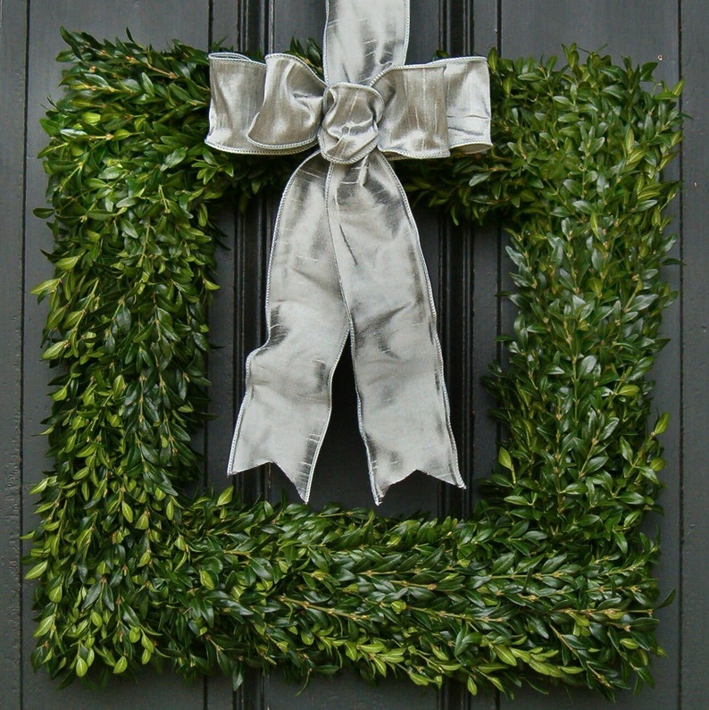 May include: A square boxwood wreath with a large silver satin bow. The wreath is hung on a black door.