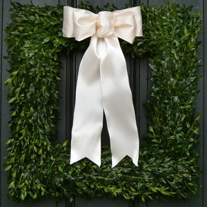 May include: A green boxwood wreath with a large white satin bow. The wreath is made of boxwood branches and is perfect for a front door or a wall.