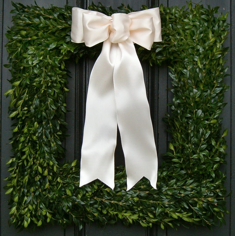 May include: A green boxwood wreath with a large white satin bow. The wreath is made of boxwood branches and is perfect for a front door or a wall.