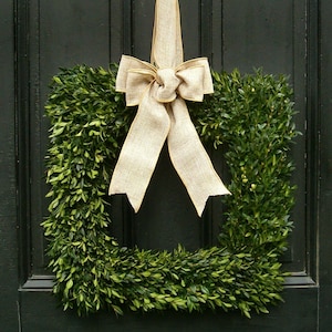 May include: A square boxwood wreath with a large burlap bow tied in the center. The wreath is hanging on a dark green door.