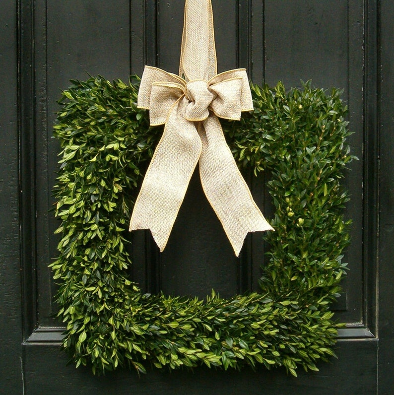 May include: A square boxwood wreath with a large burlap bow tied in the center. The wreath is hanging on a dark green door.